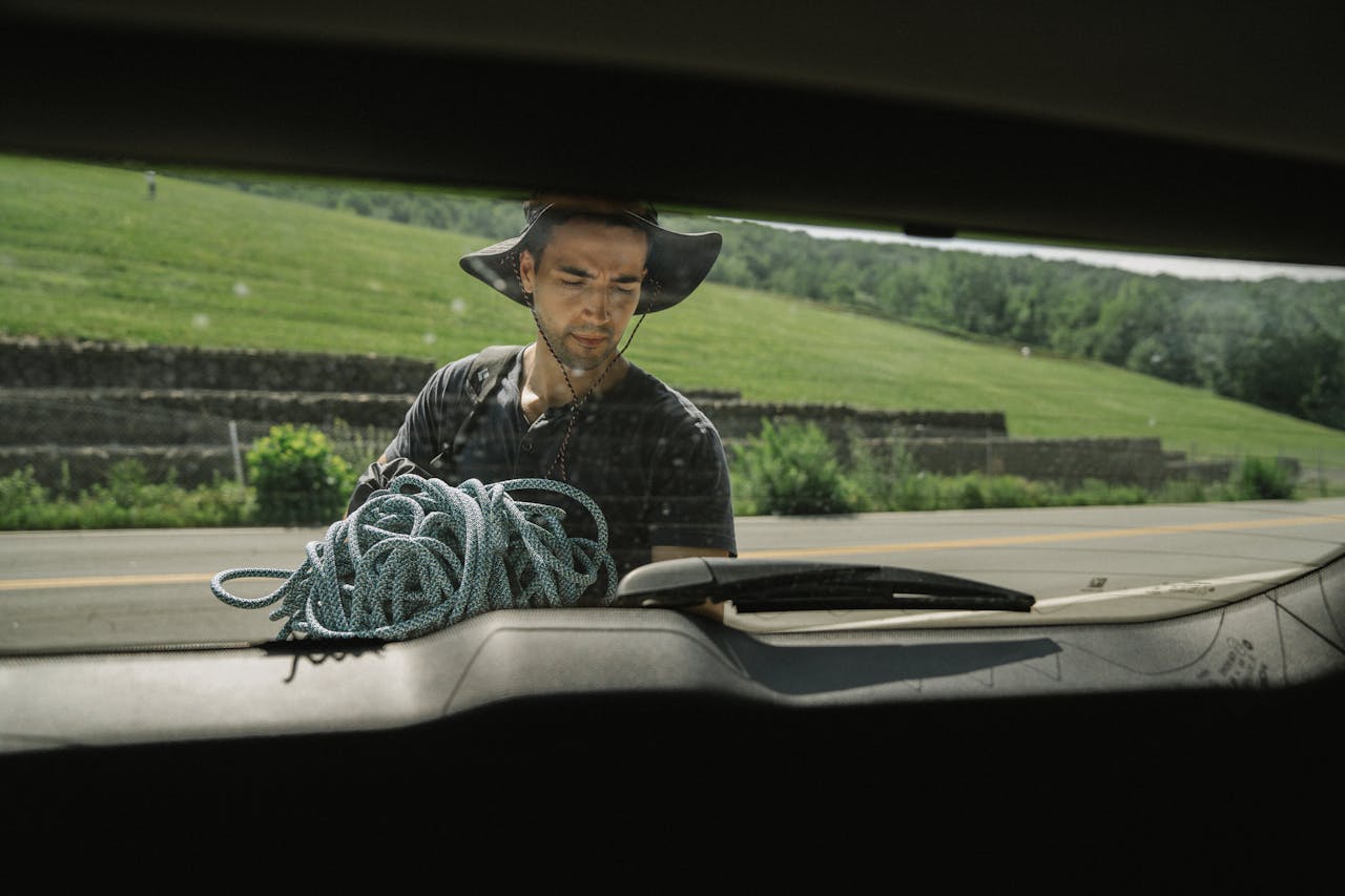 A man in a hat seen through a cars rear windshield, arranging rope during a sunny day outside.